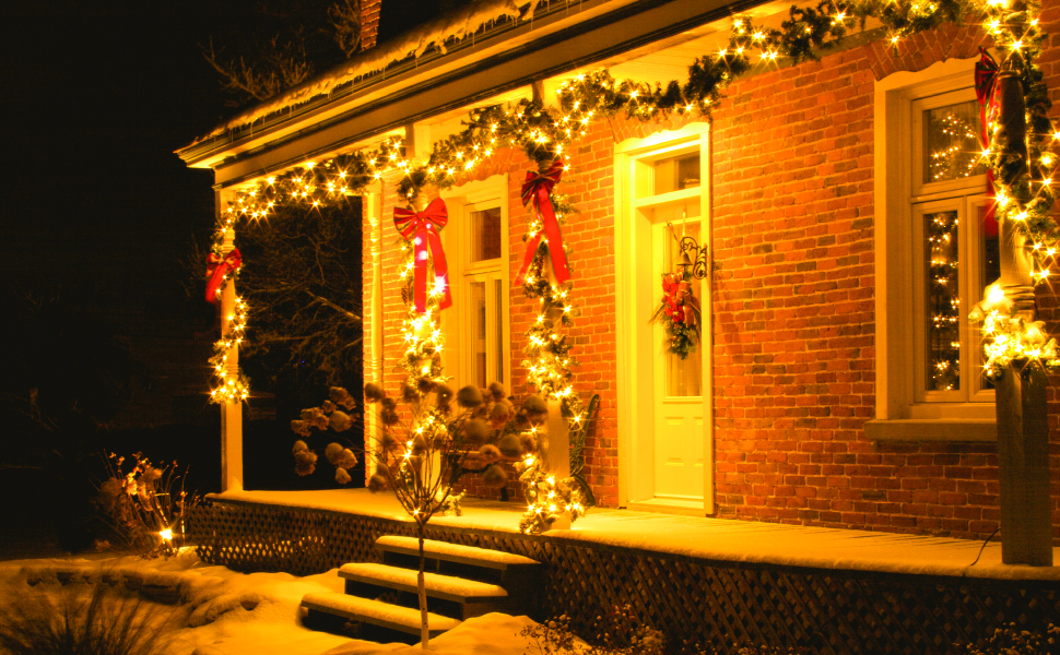 Brick house exterior decorated with warm white Christmas lights along roof, windows, and door. Features wreaths and snow-covered steps.