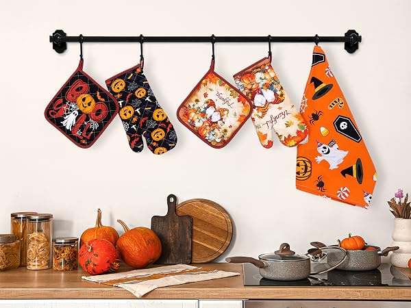 Halloween-themed kitchen accessories hanging on black rod, including oven mitts and pot holders with ghost and pumpkin patterns in orange and black.