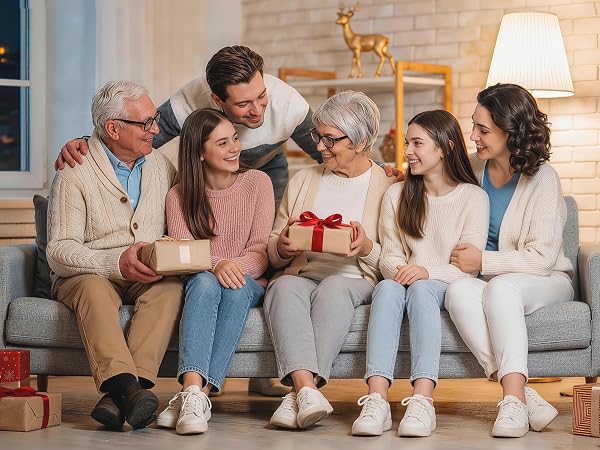Multi-generational family group sitting together on gray couch indoors, smiling and holding wrapped gift box, casual home setting with warm lighting and decorative elements.