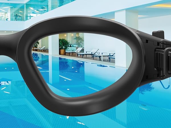 Black swimming goggles lens framing view of indoor pool. Clear water and poolside lounge chairs visible through goggle lens.