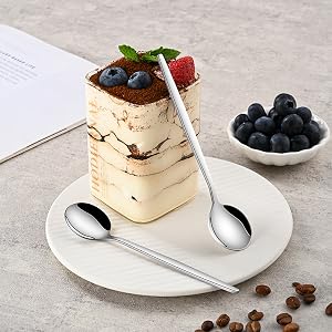 Layered dessert served on white plate with fresh blueberries and strawberry garnish. Side bowl of blueberries and coffee beans scattered on grey surface. Two silver spoons included in setting.