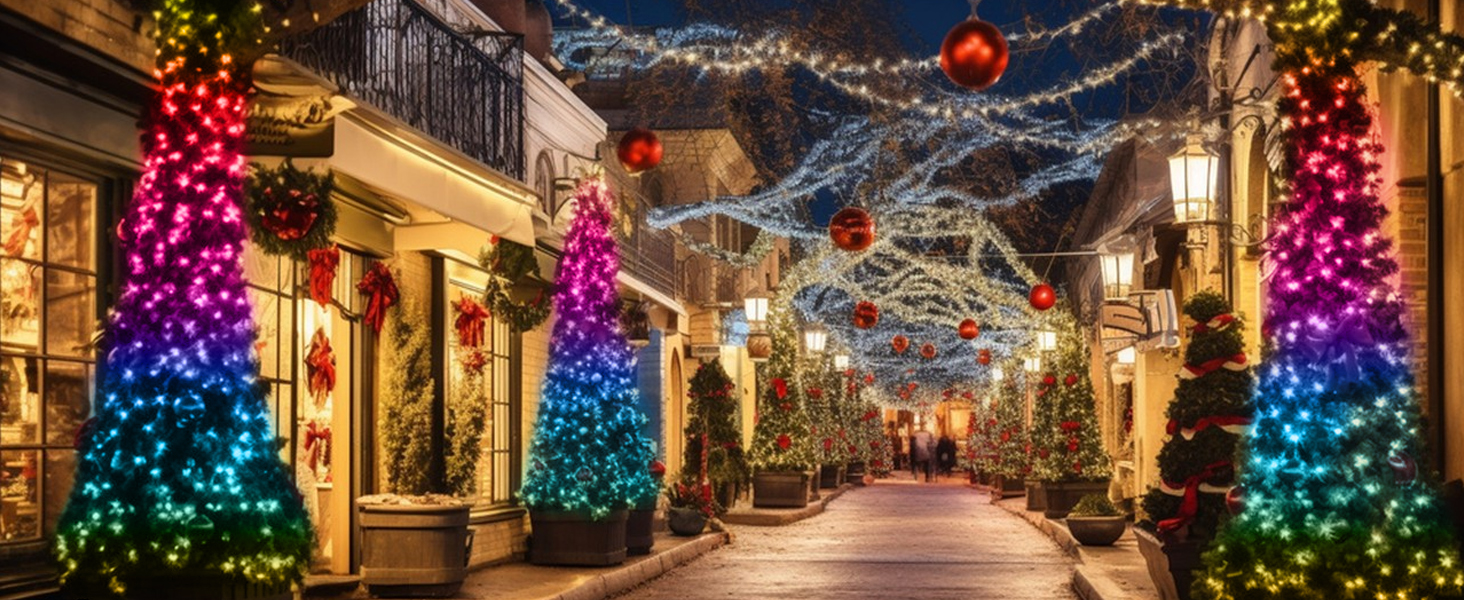 Holiday-decorated shopping street with illuminated rainbow-colored tree sculptures and hanging blue lights creating a festive walkway between storefronts.