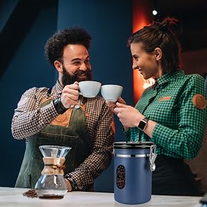 Two people in aprons sharing coffee at a counter with brewing equipment. Blue coffee container and glass coffee maker visible on the surface.