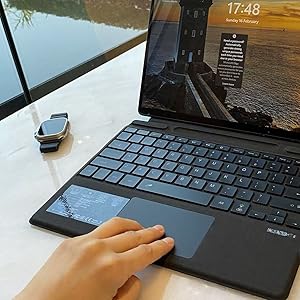 Laptop with detachable keyboard and touchpad, displaying sunset scene on screen. Smart watch visible nearby. Hand interacting with touchpad.