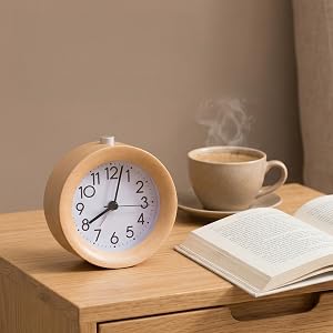 Wooden round alarm clock with blue numbers and hands displaying 10:10, positioned on wooden table beside open notebook and beige ceramic mug with steam rising from hot beverage.