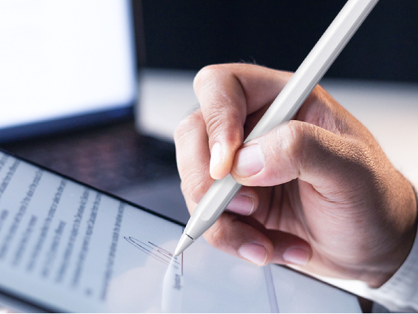 A hand holding a white stylus pen, writing or drawing on a tablet or touchscreen device with visible text.