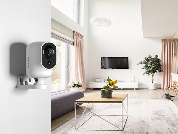 White wireless security camera mounted on wall corner in a bright, modern living room with pink curtains and minimalist furniture.