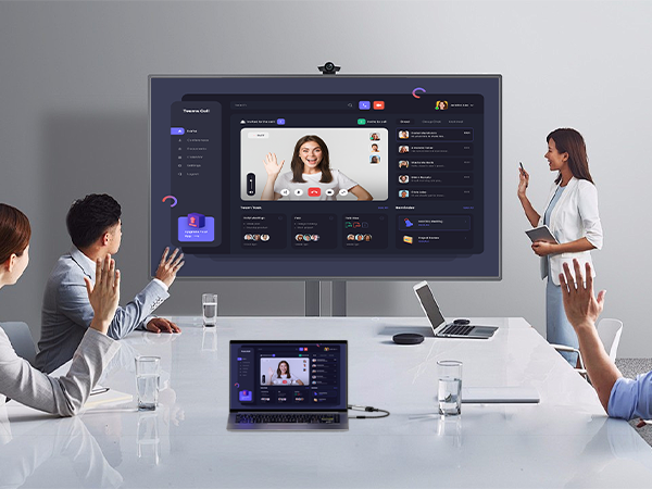 Video conferencing setup showing large wall-mounted display and laptop in conference room. Multiple people seated at table participating in virtual meeting.