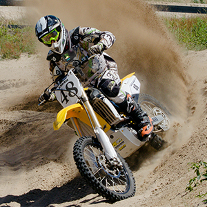 Motocross rider on dirt bike in mid-air, kicking up dust cloud during off-road racing action on sandy terrain.
