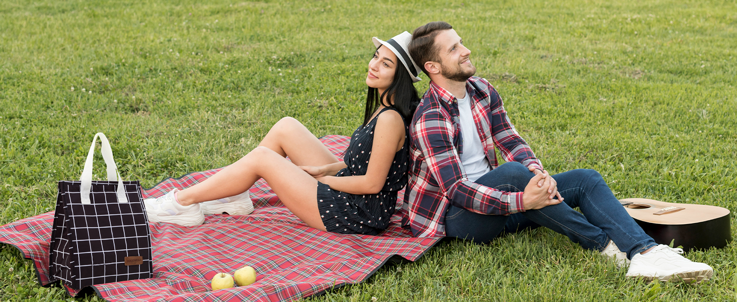 People relaxing on a red and white checkered picnic blanket on grass, with tote bag and snacks visible in outdoor setting.