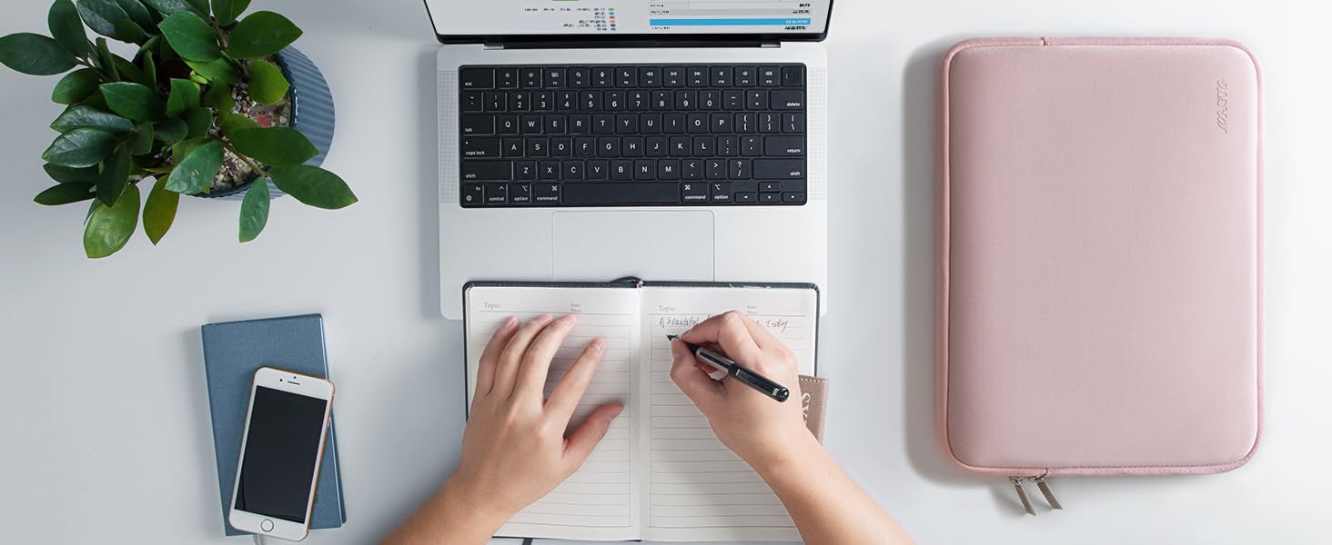 Minimalist desk setup showing laptop, pink external device, smartphone, and hands writing, with small plant decoration.