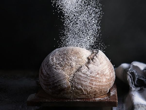 Hands sifting flour over a round loaf of bread on a dark surface, creating a cloud of fine powder.