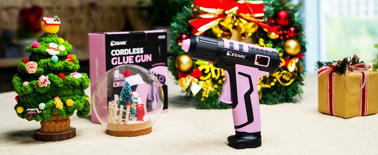 Pink cordless glue gun displayed with Christmas decorations, including miniature tree and wrapped gift against blurred festive background.