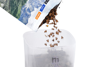 Transparent plastic container with brown pellets falling into it from a blue and white package. Container marked with 'M1' label.