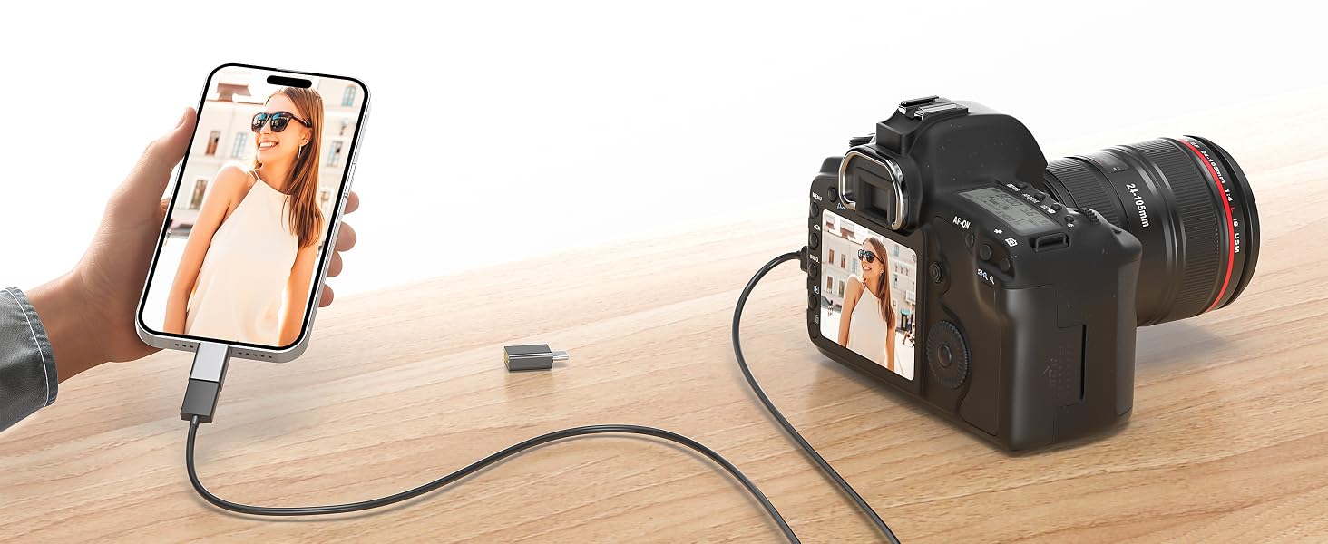 Hand holding smartphone displaying image connected via cable to black camera on wooden surface with dark background.