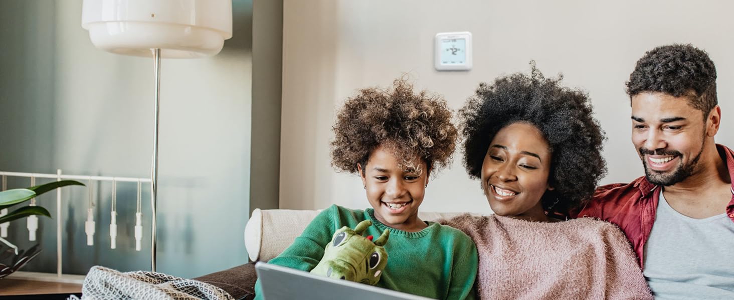 Family sitting on couch together using laptop computer, thermostat installed on background wall