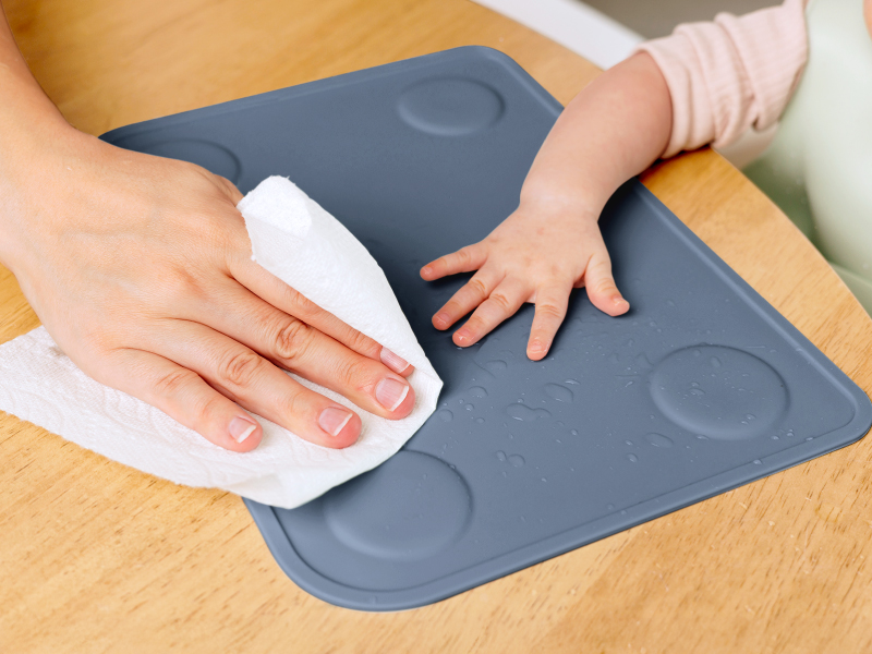 Parent wiping Prep silicone placemat clean after meals with baby hand nearby on dining table
