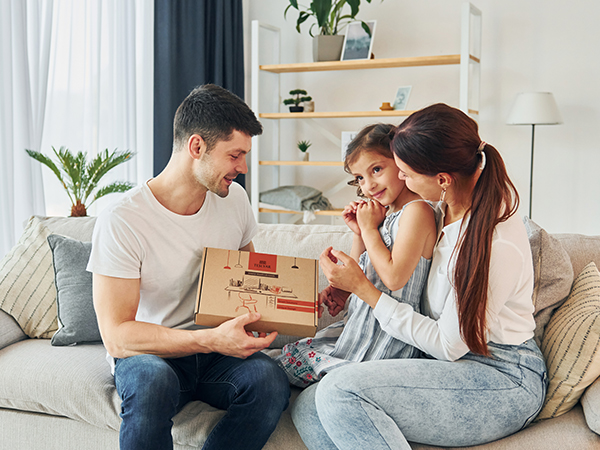 Family on couch: man, woman, and young girl examining a cardboard box together in a living room with plants and shelves.