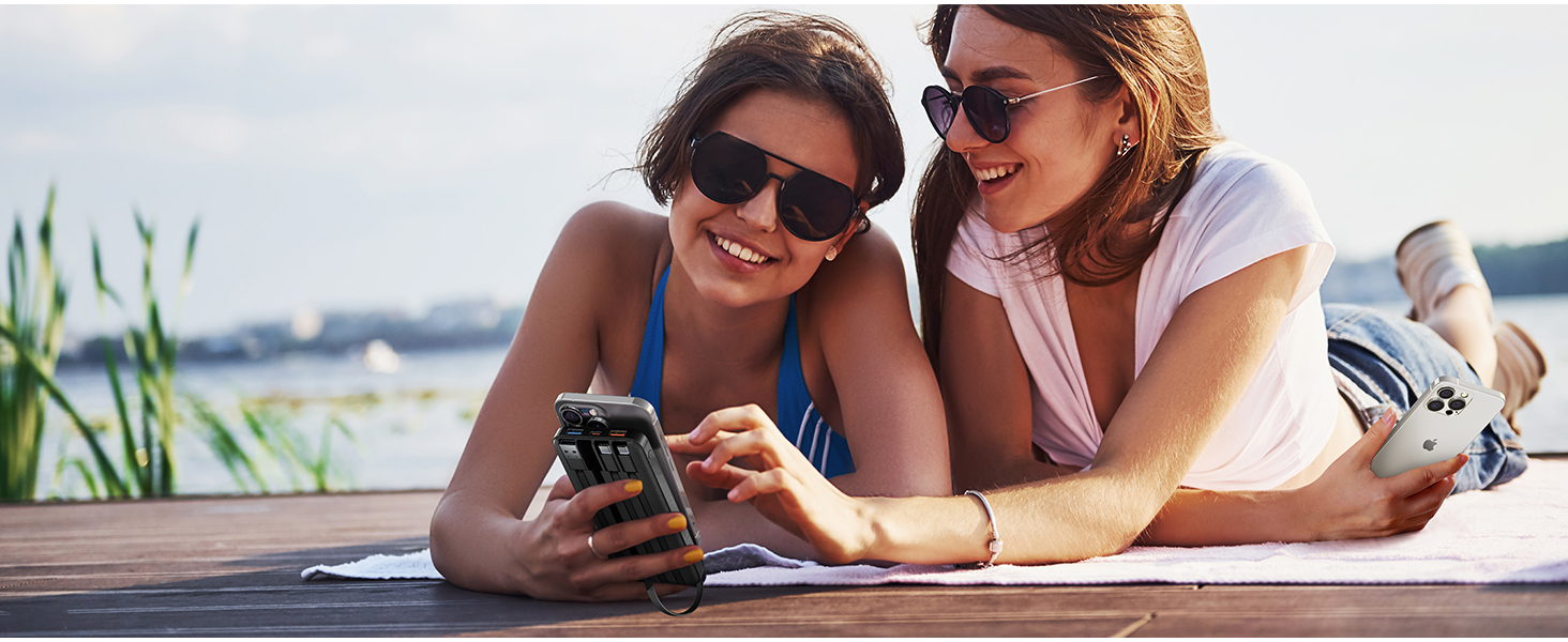 Two people wearing sunglasses, lying on a wooden surface near water, looking at a smartphone together.
