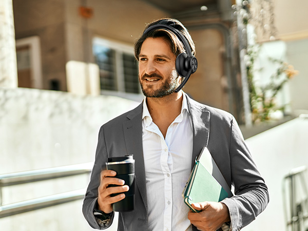 Professional wearing over-ear headphones and grey suit jacket, holding mobile device and folder while standing outdoors.