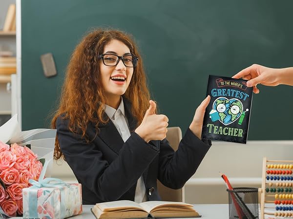 Smiling teacher sits by a bouquet of roses as a student hands her a thank you card for teacher.