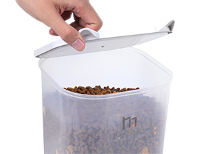 Hand opening a large transparent plastic container filled with brown pet food. Container has a hinged white lid and measurement markings on the side.