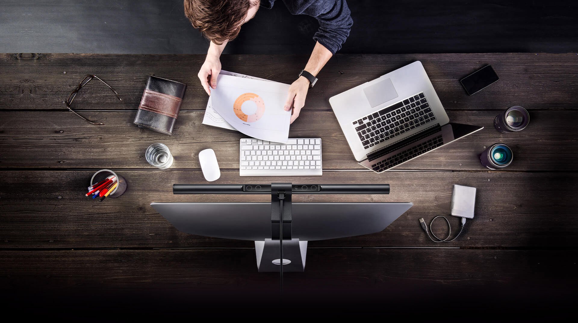 overhead view of the BenQ ScreenBar on top of an imac with a man working on a desk that has a cup of pens, glasses, leather-bound notebook, cup of water, mouse, keyboard, open laptop, smartphone, camera lenses and external hard drive with cable