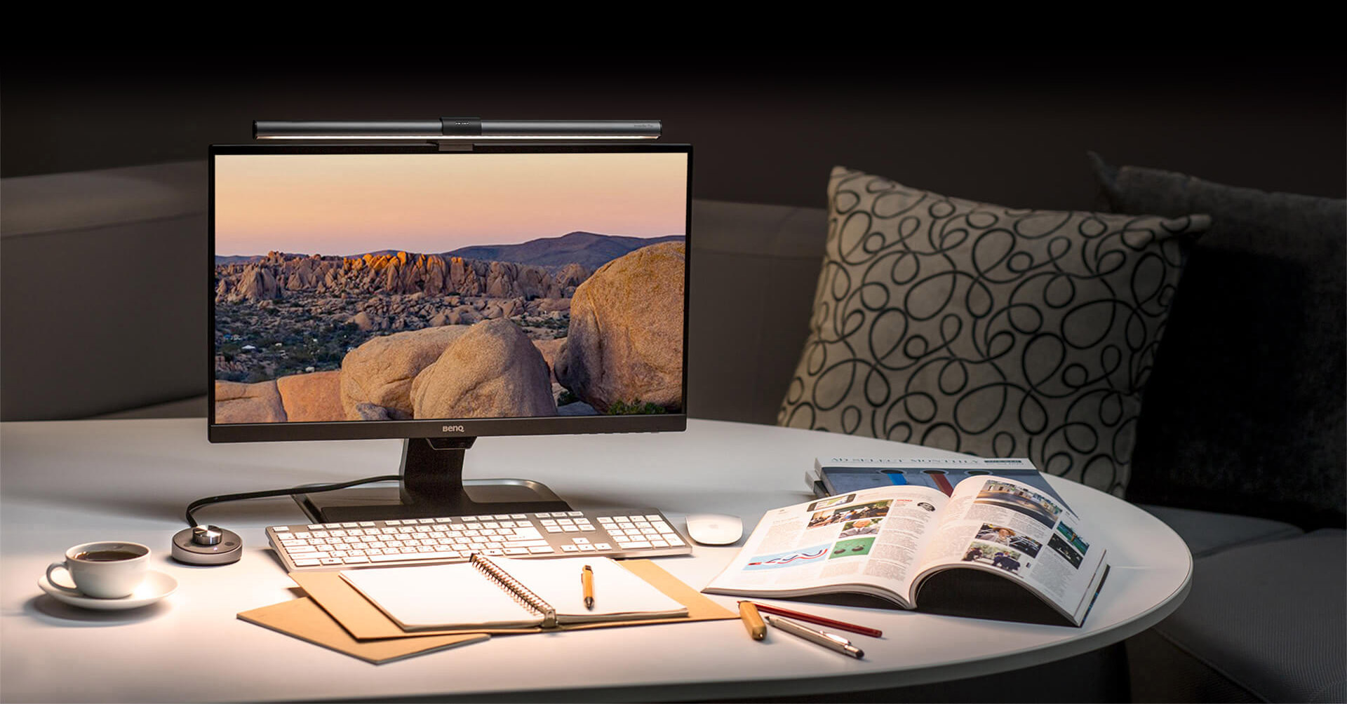 BenQ ScreenBar Plus on a monitor that's standing on a living room coffee table in front of a couch. The monitor is showing rocky plateau at dusk. On the coffee table are a cup of coffee, open notebook, pencils, the desktop dial and an open textbook