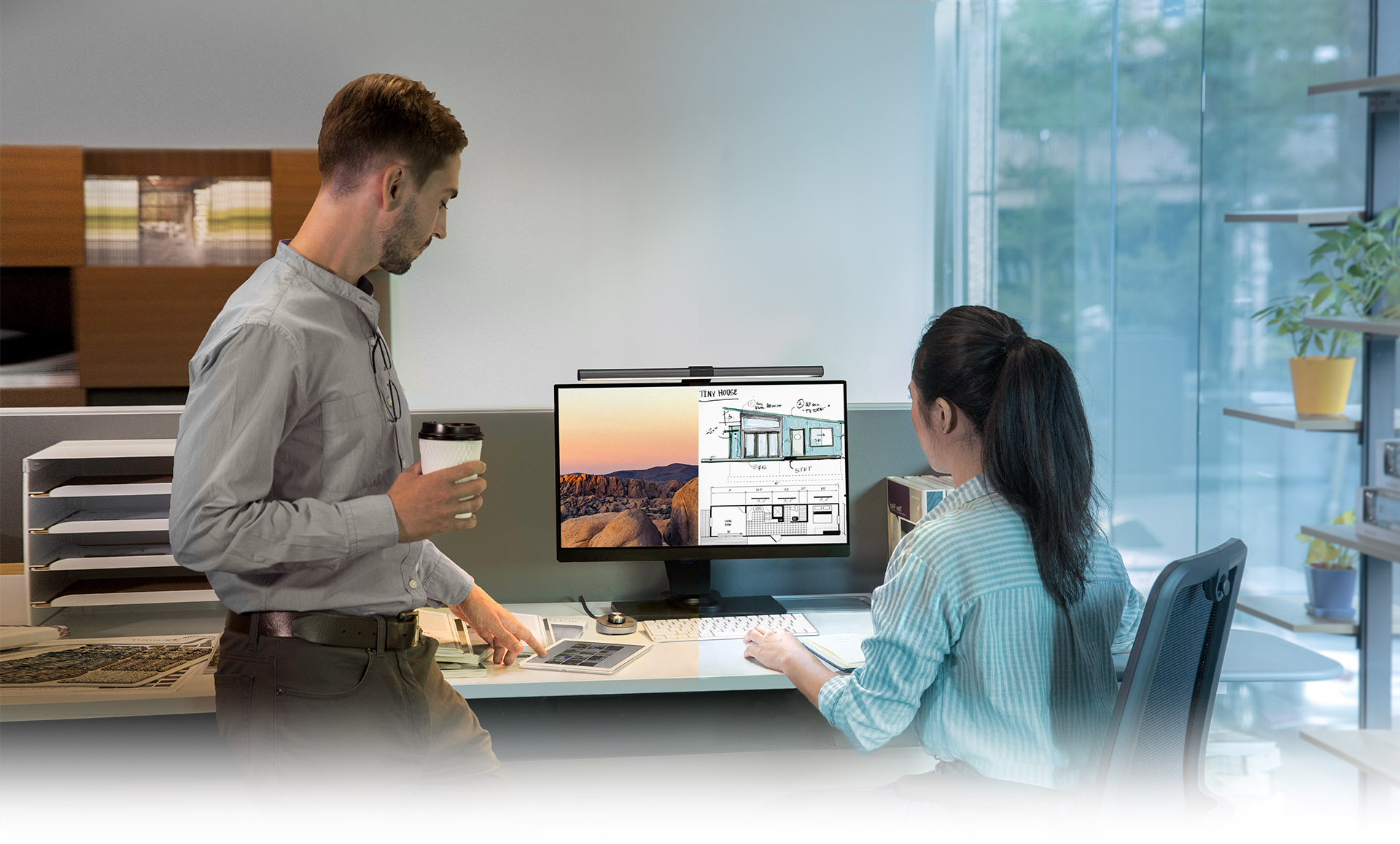 A male coworker pointing to a tablet on a female coworkers desk. She is looking at a split monitor with the BenQ ScreenBar Plus mounted on top