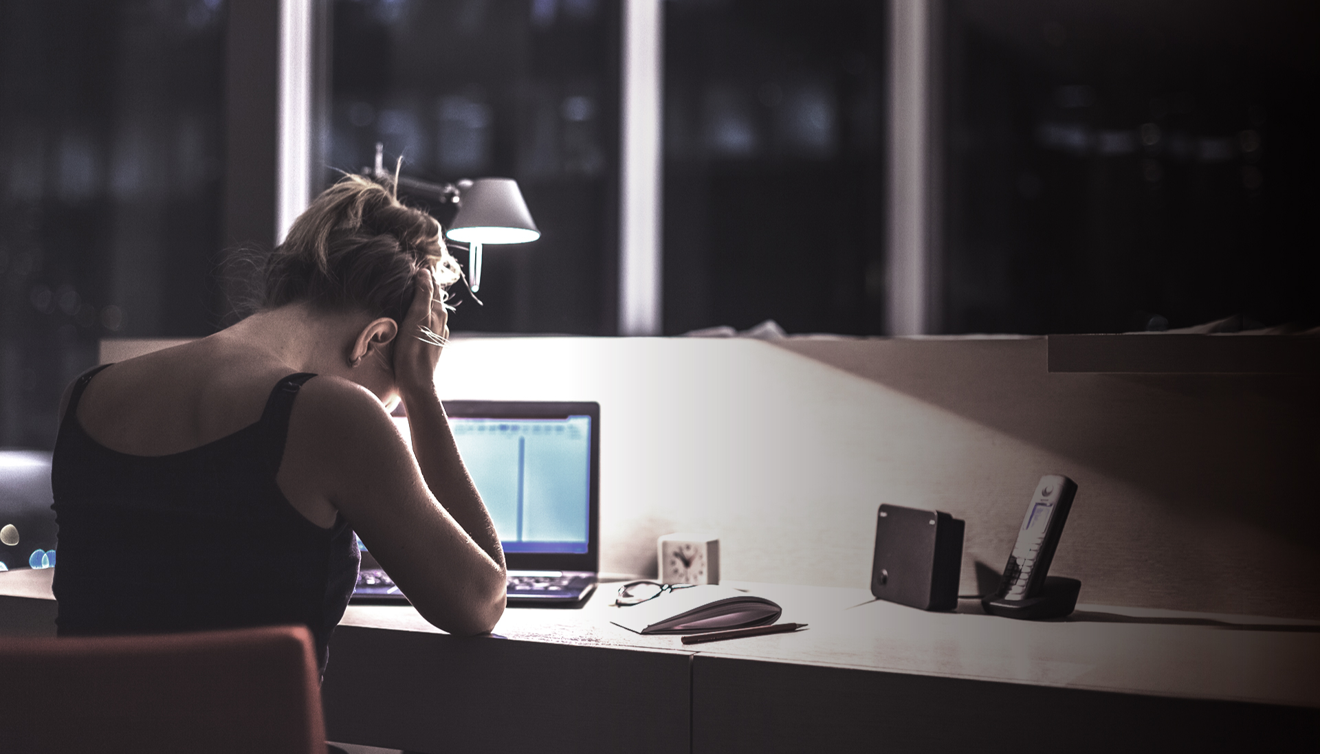 A woman with her hands on her head as she looks down tired at her desk in front of an open laptop