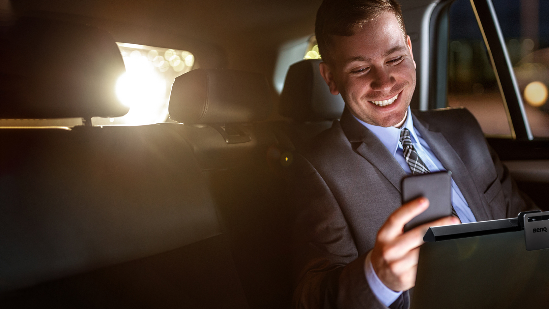 A man smiling at his phone in the backseat of a car with the BenQ ScreenBar Lite mounted in front him