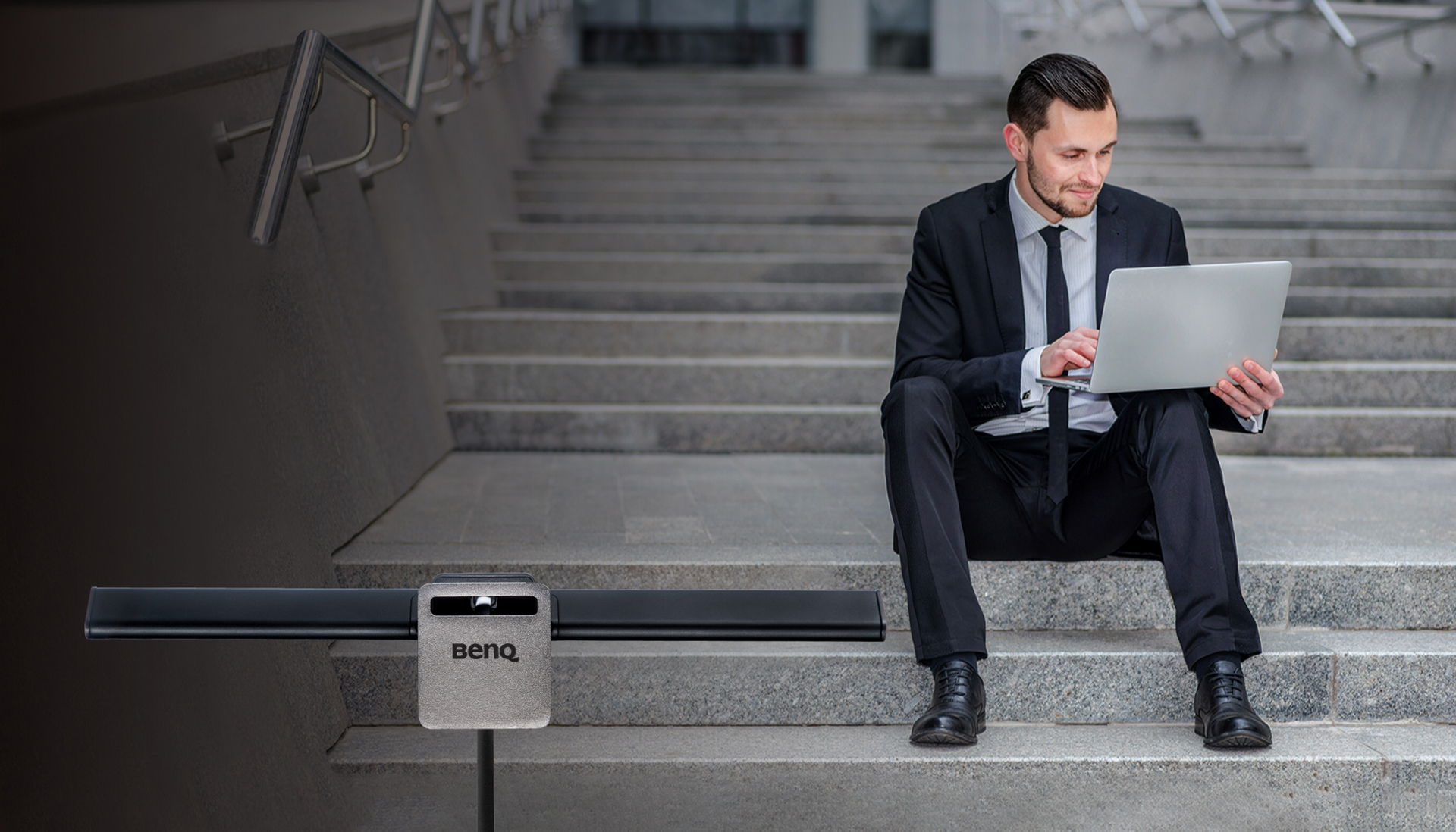 BenQ ScreenBar Lite facing forward in front of an image of a man smirking at his open laptop as he sits on concrete steps