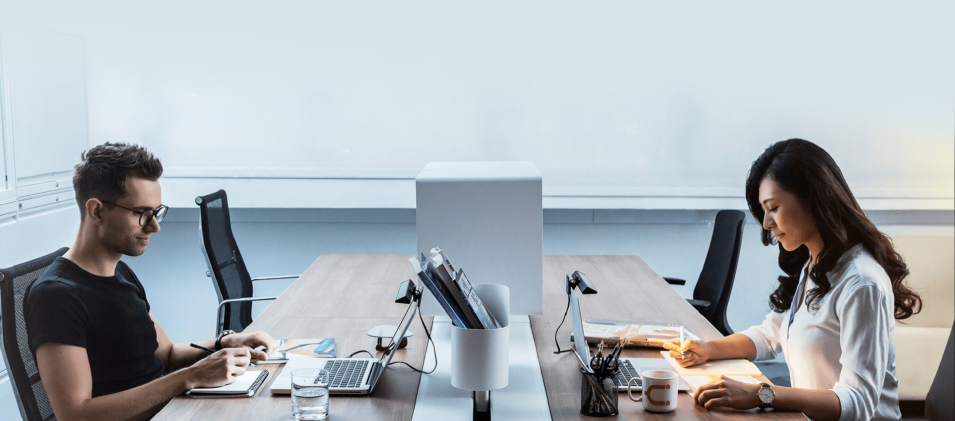 Two office workers at a conference room table sitting and taking notes in front of their open laptops that both have mounted BenQ ScreenBar Lite devices