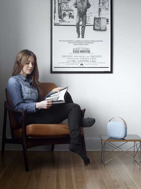Blue Helsinki Portable Speaker on a table next to a seated woman reading a magazine