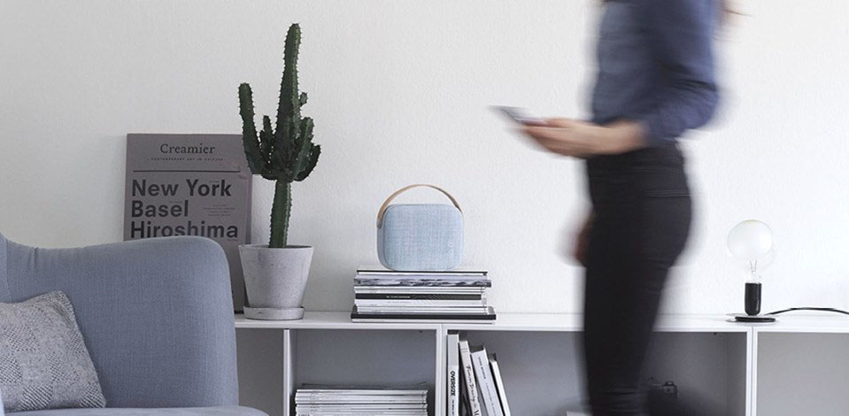 Blue Helsinki SPeaker on a Stack of Records on a Home Shelf with a Person on Their Phone Walking By