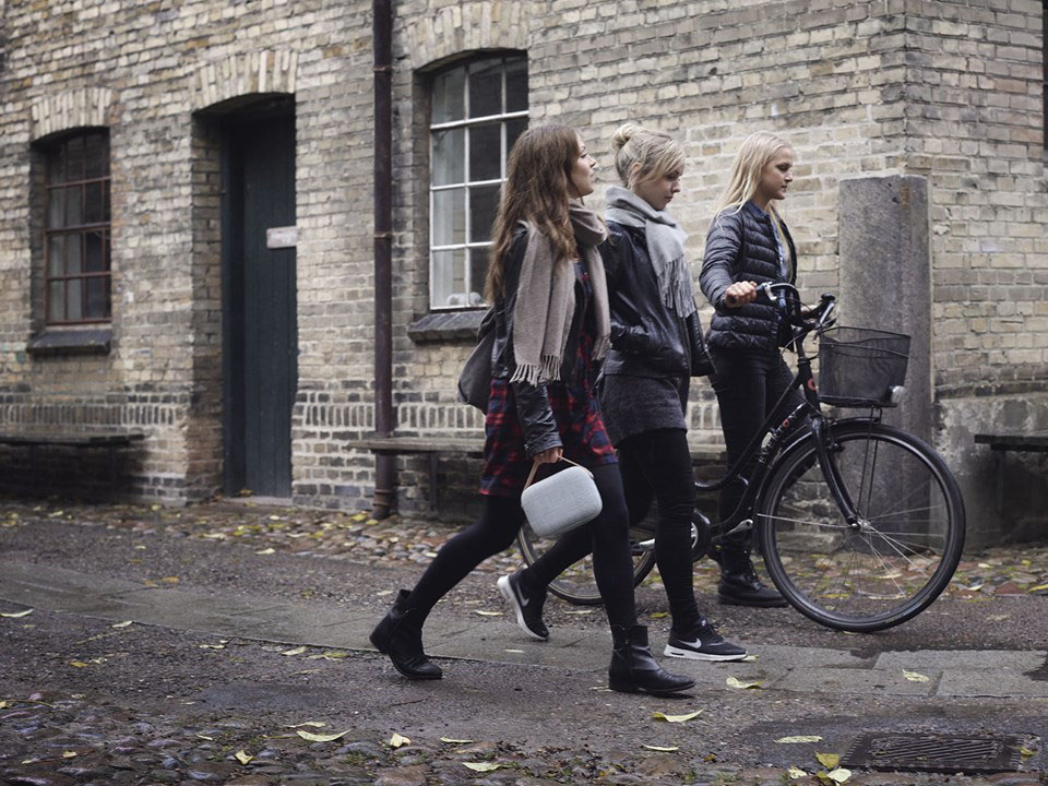Three Women in Winter Attire Walking Outside Next to a Brick Building, the woman in front carries a gray Helsinki speaker