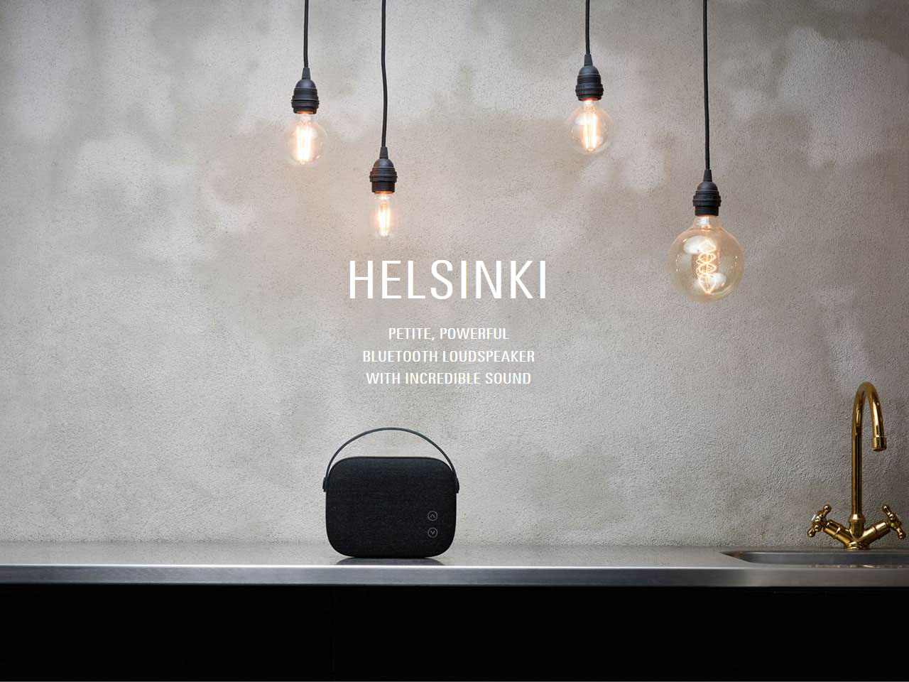 Vifa Helsinki Wireless Portable Speaker on a Metal Kitchen Counter Next to a Sink, Below Dangling Lightbulbs