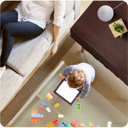    A boy watching a tablet. Behind him is a Deco M5 placed on a wood desk. Next to him on the right is her mother lying on a couch