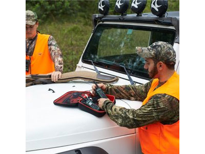  A man standing by an SUV, operating a radio stored in a case, with another radio stored in the case. On the other side of the car is another man holding some hunting gear in his hand  