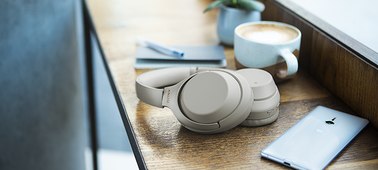    The Sony WH-1000XM3 on a desk behind a window, with a mobile phone, a potted plant, and a notebook with a pen on it
