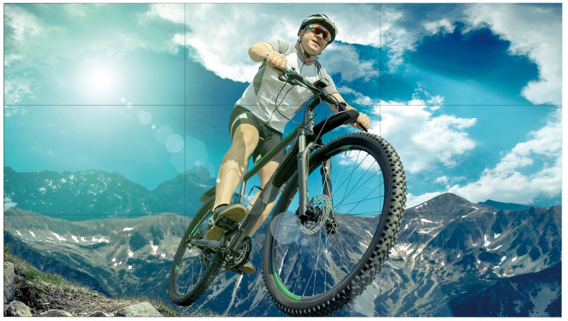 LG display showing a man mountain biking with the sun shining behind him and multiple mountains on a cloudy blue sky behind him