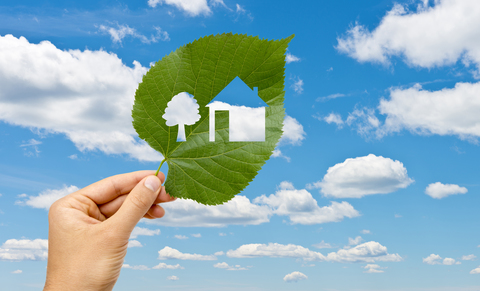 A hand holding a leaf that has a tree and house shape cut of it. The background is a blue sky with white clouds