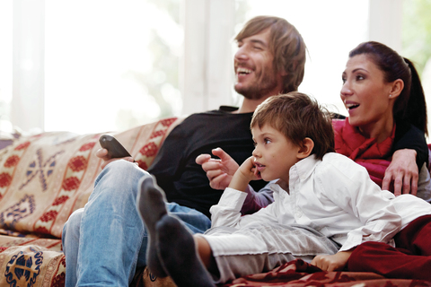 A family of three (father, mother and son) on a couch laughing. The father holds a remote.