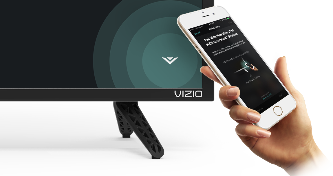 Hand holding up a smartphone angled up to the left with the VIZIO smartcast app open for setup. Behind the smartphone is the bottom right corner of a VIZIO TV