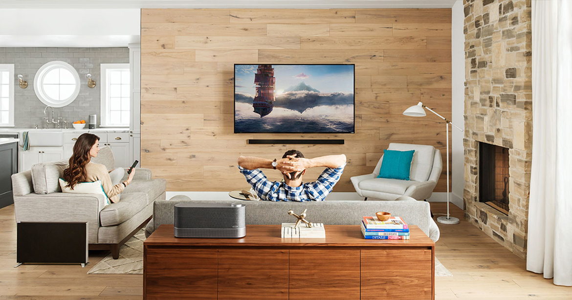 Vizio E43u-D2 TV mounted on a living room wall above a mounted speaker. The living room has a man and woman lounging on their couches