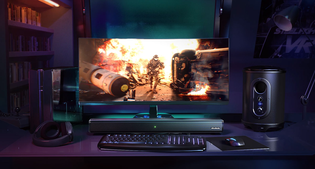 AverMedia Soundbar Under a Monitor with Fiery War Gameplay, in Front of a Keyboard and Mouse on a Mousepad, Next to a Subwoofer, a Shelf of books and desk lamp