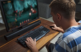 A Male Gamer Playing on his wooden desk with monitor, keyboard, mouse and AverMedia soundbar