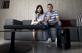 A man and woman watching TV on a couch with the Avermedia soundbar and subwoofer on the floor and coffee table in front of them