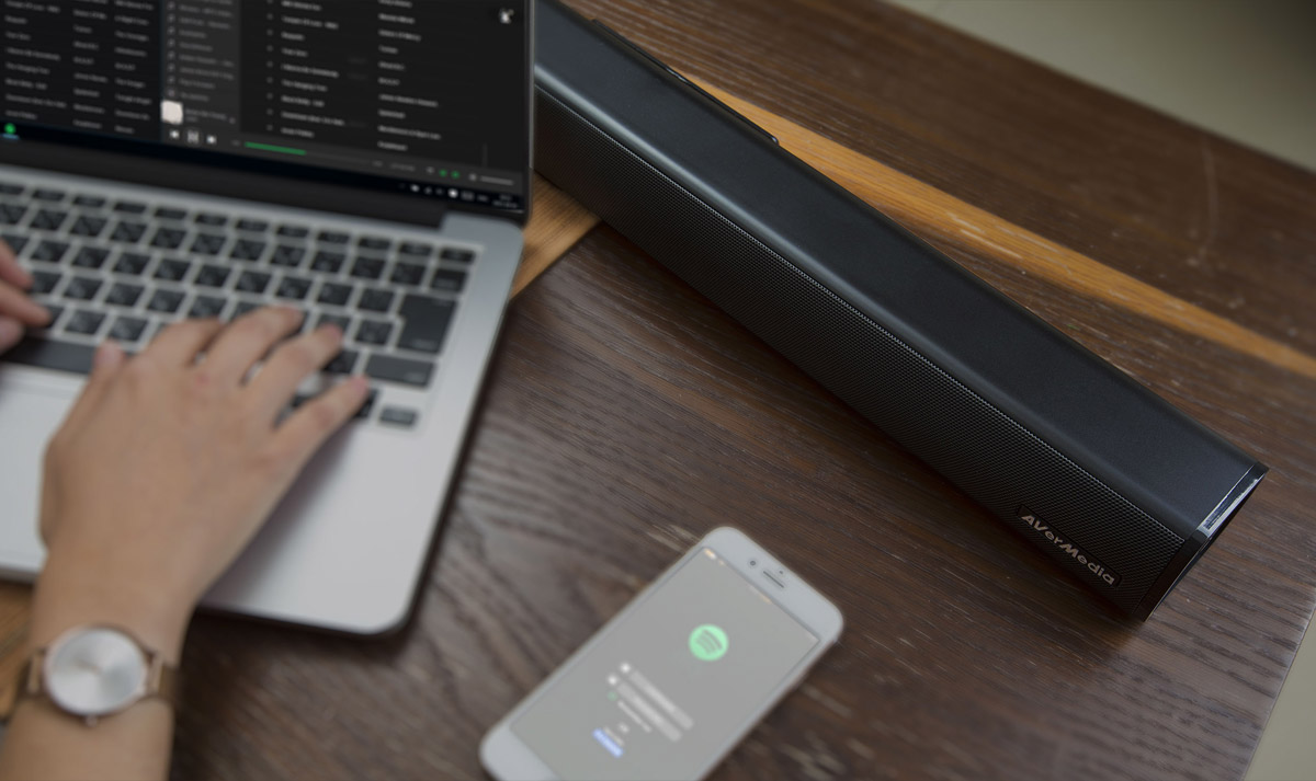 AverMedia Soundbar on a desk next to a person using an open laptop with spotify and smartphone that is also connected to spotify