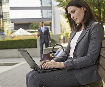 A woman in office attire outside, sitting on a bench with her laptop open as she types